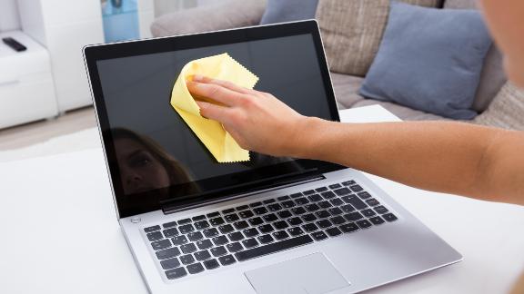 A person cleaning a laptop screen with a yellow microfiber cloth. The laptop is placed on a white table in a living room with a grey couch and cushions in the background.